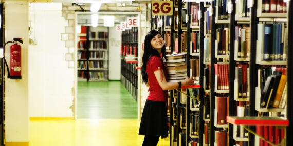 View of a library with bookshelfes. A young woman is carrying a pile of books. 